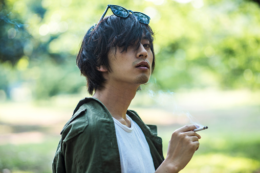 Portrait of a young Japanese man in Tokyo, Japan. He is serious, smoking cigaret and is thoughtful. Green trees in the back.