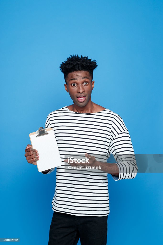 Surprised afro american holding clipboard Surprised afro american young man wearing striped top holding clipboard in hand. Studio portrait, blue background. Adult Stock Photo