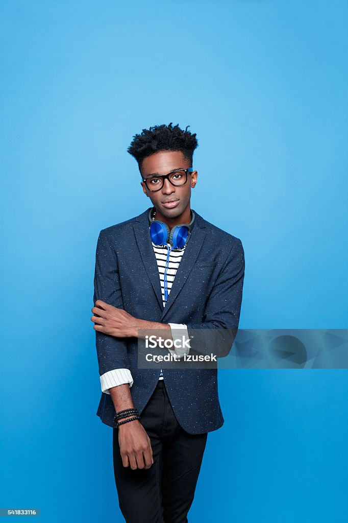 Afro american guy in fashionable outfit Fashionable afro american young man wearing striped top, navy blue jacket, nerd glasses and headphone, looking at camera. Studio portrait, blue background. African Ethnicity Stock Photo