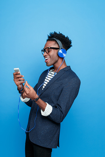 Side view of fashionable afro american young man wearing striped top, navy blue jacket, nerd glasses and headphone, using a smart phone, listening to the music. Studio portrait, blue background.