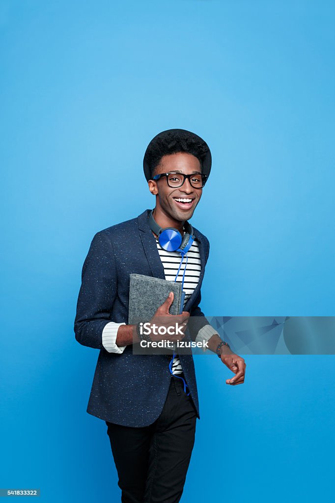 Excited afro american guy in fashionable outfit, holding notebook Studio portrait of happy afro american young man wearing striped top, navy blue jacket, hat, nerd glasses and headphone, smiling at camera, holding a notebook in hand. Studio portrait, blue background. Colored Background Stock Photo