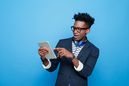 Studio portrait of surprised afro american young man wearing striped top, navy blue jacket, nerd glasses and headphone, holding a digital tablet in hand. Studio portrait, blue background.