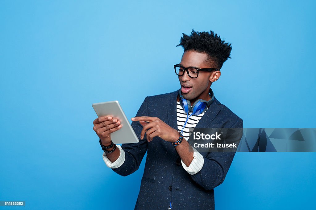Excited afro american guy in fashionable outfit, holding digital tablet Studio portrait of surprised afro american young man wearing striped top, navy blue jacket, nerd glasses and headphone, holding a digital tablet in hand. Studio portrait, blue background. Men Stock Photo