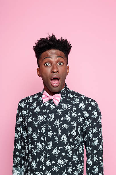 Surprised afro american young man Portrait of surprised afro american young man wearing floral pattern shirt and pink bow tie, staring at cmaera with mouth open, rolling his eyes. Studio shot, pink background. gasping stock pictures, royalty-free photos & images