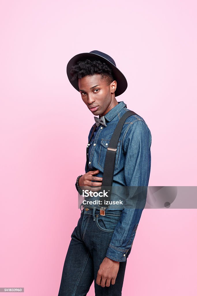 Fashionable afro american nerd guy Portrait of confident afro american young man wearing denim clothes, bow tie, suspenders and hat, looking at camera. Studio shot, pink background. Fashion Stock Photo