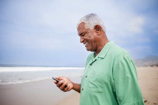 A senior African American man checks his phone on the beach.