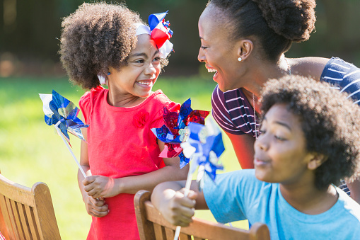 An African American mother with two mixed race children celebrating an American patriotic holiday, perhaps July 4th or Memorial Day. They are playing with red, white and blue pinwheels, smiling and laughing, outdoors on a bright, sunny day. The children are black, Asian and Hispanic. The focus is on the woman and girl who are looking at each other.