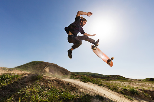 Young man exercising skateboarding on dirt road while being in Ollie position in mid air against the blue sky.  Copy space.