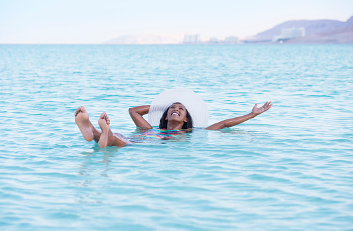 Happy woman enjoying vacation in Dead Sea. Attractive lady with white sunhat floating in salty lake.