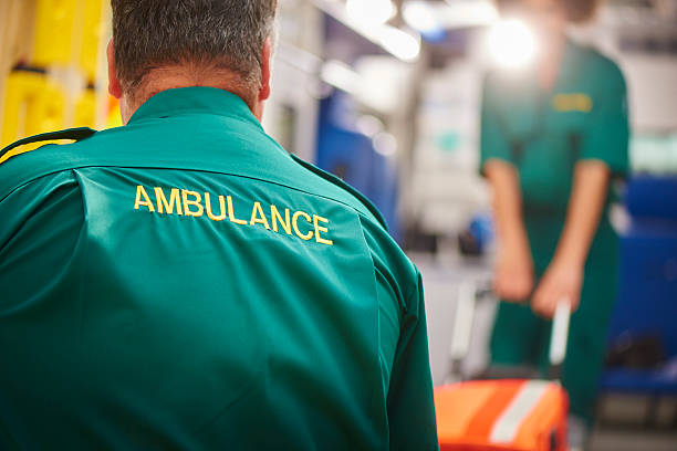 ambulance crew pulling stretcher two uk ambulance staff members pull a stretcher on or off of their ambulance . We are looking over the shoulder of the male paramedic focussing on the word ambulance on his back.They are wearing a green ambulance uniform typical of uk paramedics. The female paramedic and the back of the ambulance are defocussed . emt stock pictures, royalty-free photos & images