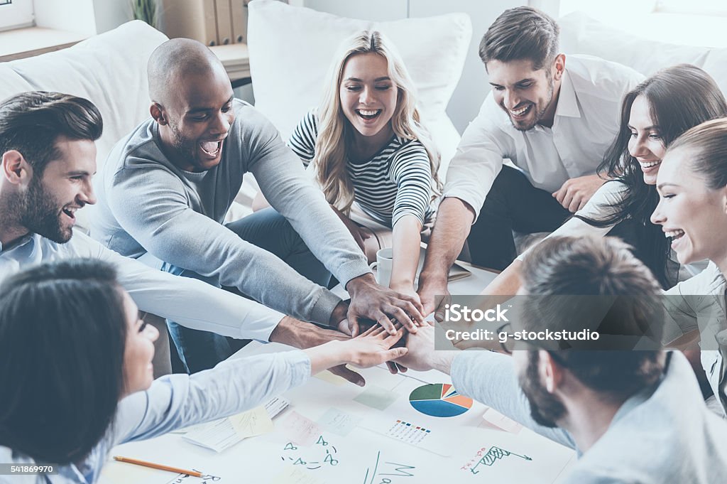 Together we are stronger! Group of happy business people holding hands together while sitting around the desk Hands Clasped Stock Photo