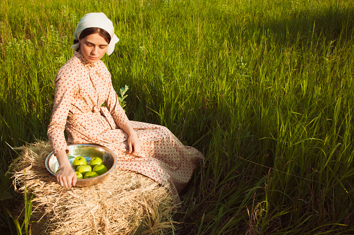 The healthy rural life. The woman in scarf sitting on a haystack with apples against green meadow
