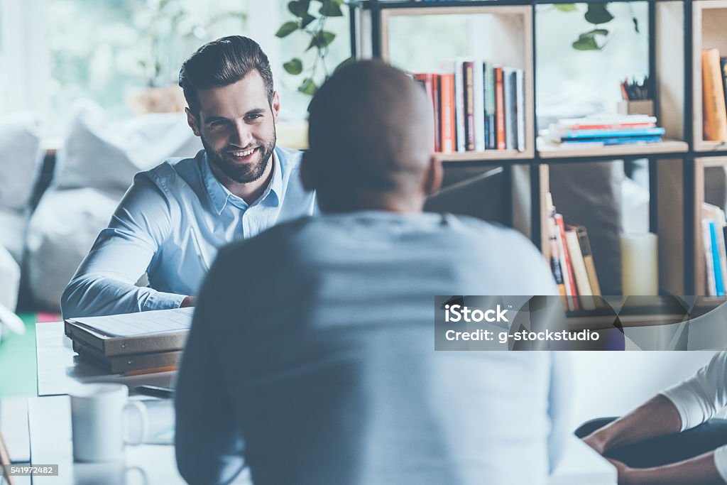 Job interview. Two young men in smart casual wear sitting at the office desk together while one of them smiling Discussion Stock Photo