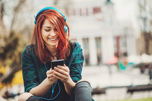 Smiling teenage girl with headphones, listening music from her smart phone outdoors at urban setting, with copy space.