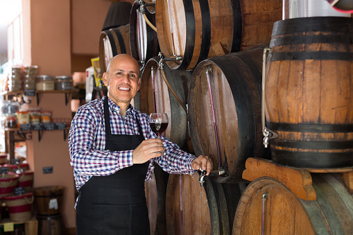 Friendly smiling mature  seller tasting wine from wood barrel  in wine house