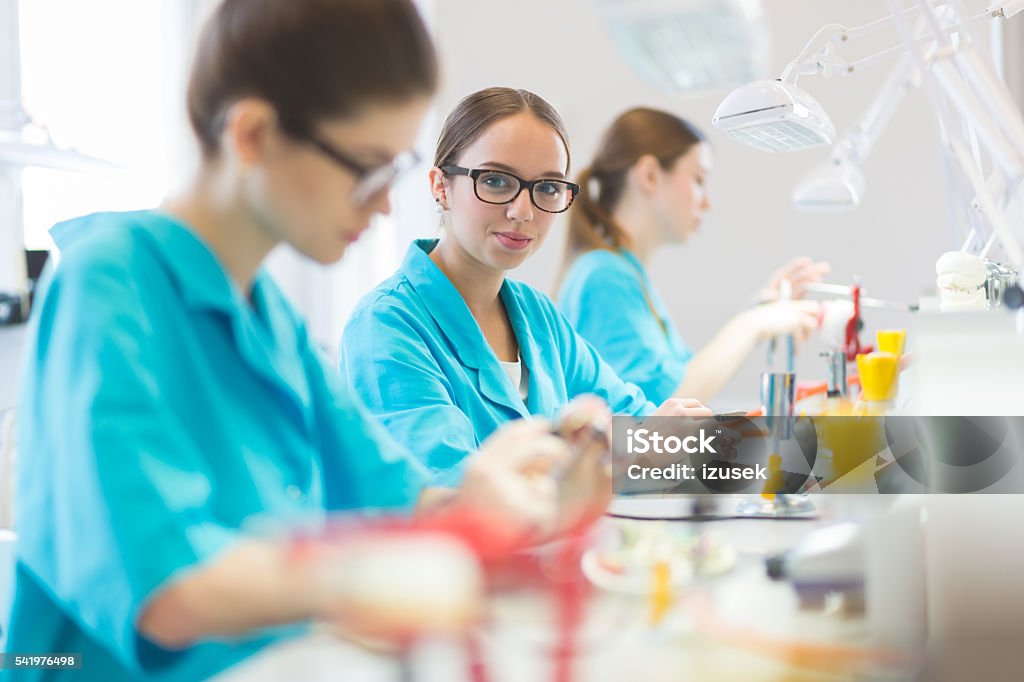 Female students learning prosthetic dentistry Female students wearing uniforms in a prosthodontic lab, learning prosthetic dentistry. Focus on friendly girl wearing glasses, smiling at camera. Adult Stock Photo