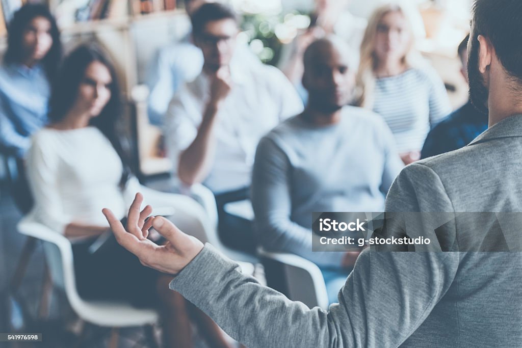 Business coach. Rear view of man gesturing with hand while standing against defocused group of people sitting at the chairs in front of him Education Training Class Stock Photo