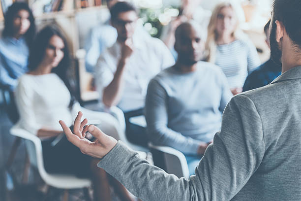 Business coach. Rear view of man gesturing with hand while standing against defocused group of people sitting at the chairs in front of him business conference photos stock pictures, royalty-free photos & images