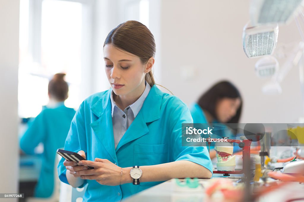 Prosthetic dentistry female student texting on smart phone Young woman learning prosthetic dentistry, sitting in a laboratory and texting on mobile phone. Dentist Stock Photo