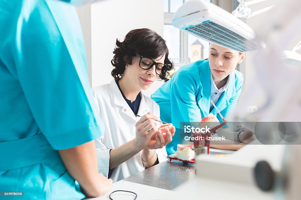 Female students learning prosthetic dentistry Female students wearing uniforms sitting in a prosthodontic lab, learning prosthetic dentistry. Focus on their teacher holding dentures. Dental Health Stock Photo