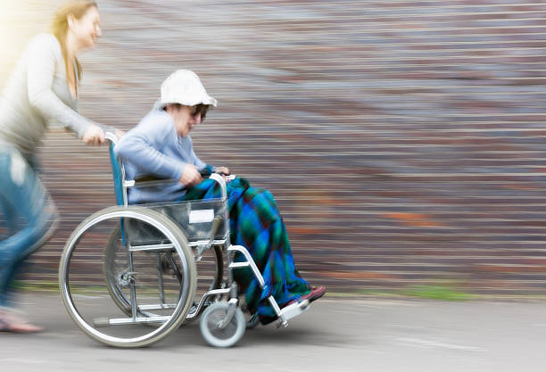 A laughing younger woman pushes an excited, smiling old woman in a wheelchair along at speed, showing motion blur, as they have some fun together. Copy space on the defocussed wall.