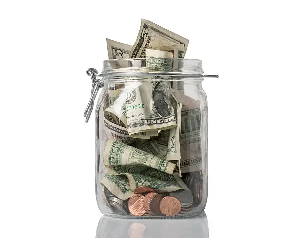 A tip jar or jar for savings filled over the top with American coins and bills.