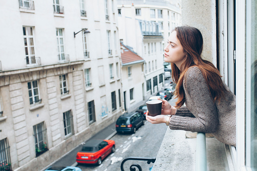 Young woman leaning on window and drinking coffee, looking away. Street view, car and building exterior on background.