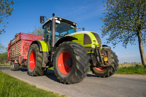 Tractor with trailer on road under clear sky. Taken in springtime in Friesland, Lower Saxony, Germany, Europe. Blurred motion and low angle view.
