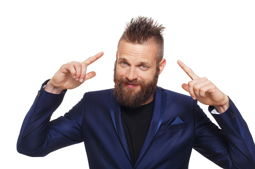 Young bearded man isolated at white background. Close up portrait of guy with beard showing, pointing fingers at his cool hairstyle, looking at camera. Boy style, trendy hipster in blue suit.