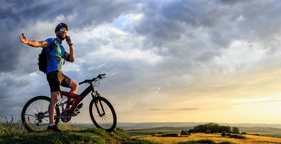 Young male mountainbiker talking on the phone on the top of the mountain. Wears sportswear, arm band, headset, sunglasses, backpack and helmet. Landscape view on background. Low angle view.