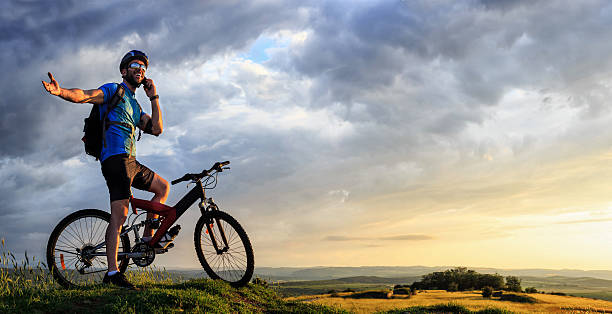 Mountainbiker using phone on top of the mountain Young male mountainbiker talking on the phone on the top of the mountain. Wears sportswear, arm band, headset, sunglasses, backpack and helmet. Landscape view on background. Low angle view. extreme phone stock pictures, royalty-free photos & images