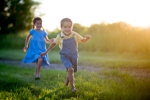 An adorable brother and sister are playing together outside on a sunny summer evening in a grassy field. They are chasing each other and playing tag.