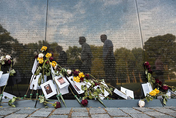 Washington DC, USA - June 20, 2016: Roses and memorial cards of the deceased are leaned against the wall at the Vietnam Veterans Memorial in Washington DC. Visitors to the memorial are reflected in the stone.