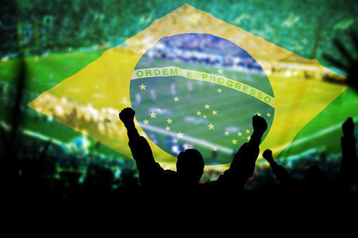 Image of a man celebrating at a sports event with his arms raised, in silhouette. Brazil flag overlayed.