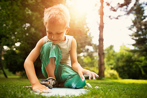 Little boy chalking on the garden stepping timber stone. The boy aged 6 is looking down at the stone. Garden and lawn visible in the background.