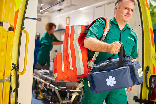 a male uk paramedic emerges from the back of his ambulance carrying a responders backpack , and vital signs monitor . Behind him a female paramedic is preparing the monitor for the arrival of casualties .