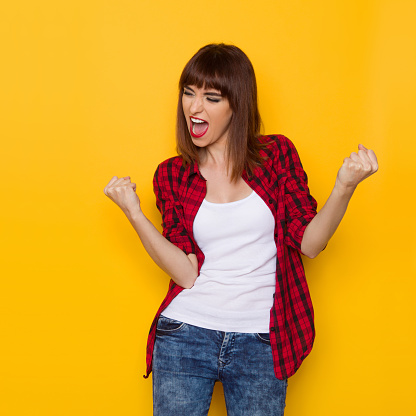 Young woman in red lumberjack shirt and jeans shouting clenches fists and looking away. Three quarter length studio shot on yellow background.
