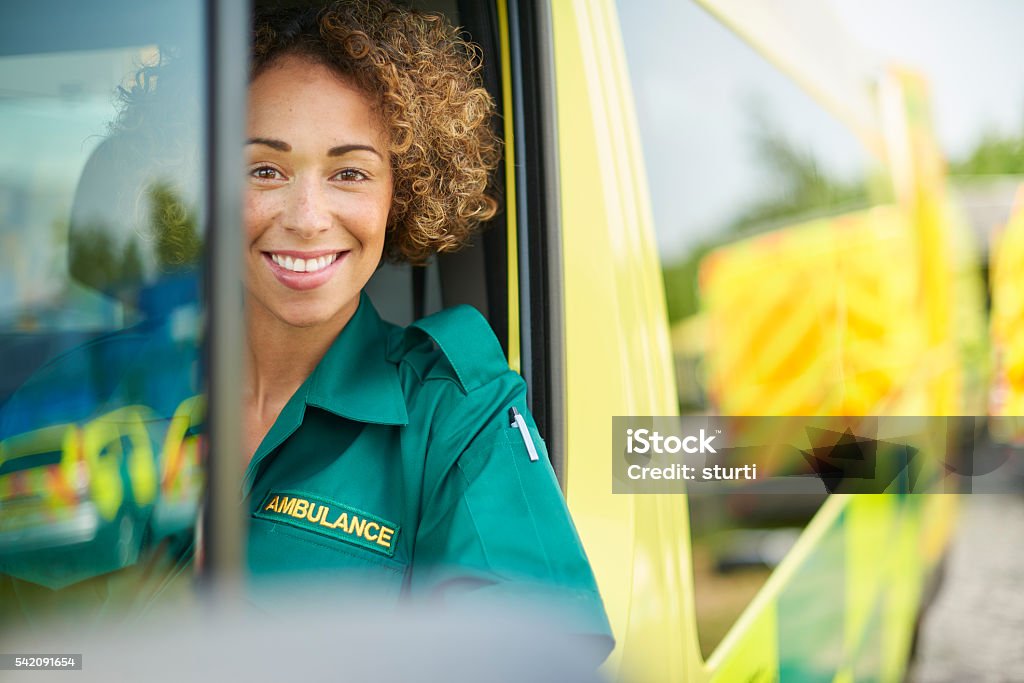 happy female paramedic uk ambulance staff members in her ambulance. She is wearing  green ambulance uniform typical of uk paramedics. She is sitting in the ambulance ready to go to a call Ambulance Stock Photo