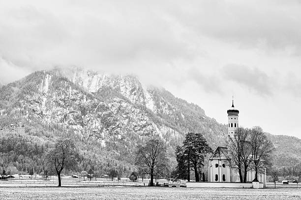 Old church with high bell tower and graveyard bellow mountains.  April weather. Wet spring snow in already fresh green grass  of meadow bellow mountains.