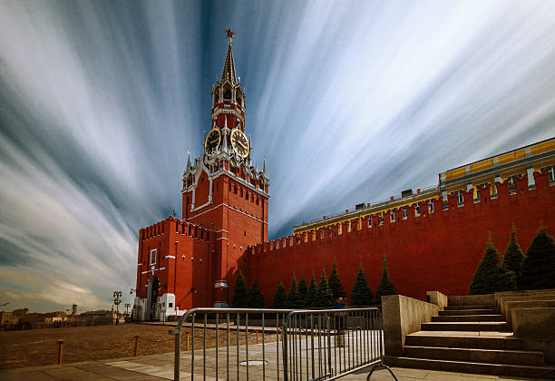 The Spasskaya Tower of the Moscow Kremlin, featuring its iconic clock face and red star, stands against a cloudy sky