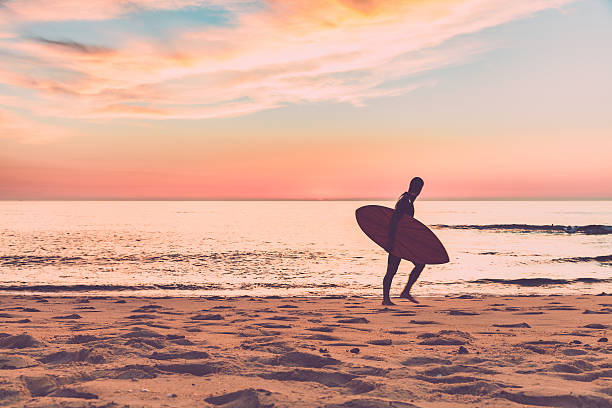 surfista caminhando na praia com uma prancha de surf ao anoitecer - surfing surfboard summer heat - fotografias e filmes do acervo