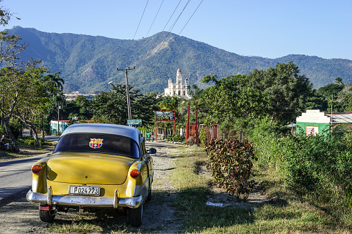 Trinidad, Cuba - January 12, 2016: The road to the Shrine Basilica Maria la Virgen della Caridad del Cobre. In the foreground, holding an old American antique car and in the background is the pilgrimage site of the Lady of Charity of El Cobre in Santiago de Cuba. It is destination of pilgrimage by thousands of believers who come daily to honor the virgin and beg her favors and blessings.