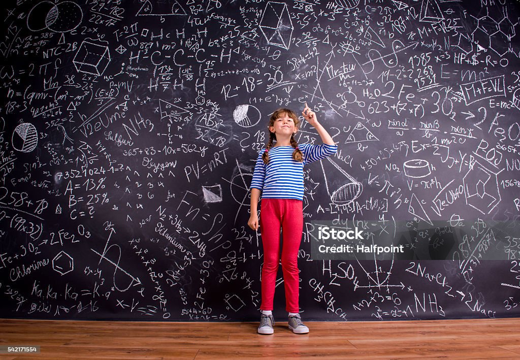 Girl with two braids, big blackboard with mathematical symbols Girl in blue striped t-shirt and red trousers, with two braids, arm raised, finger raised, against big blackboard with mathematical symbols and formulas Child Stock Photo