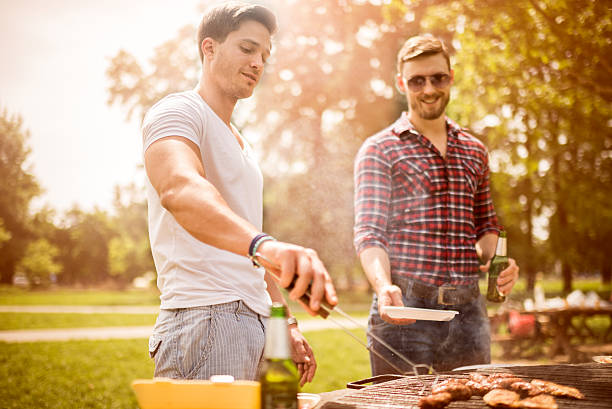 Male friends on a barbecue picnic Guys having fun during barbecue picnic in the park or back yard. One of them is holding a plate and beer while the other is cooking and serving. roast beef photos stock pictures, royalty-free photos & images