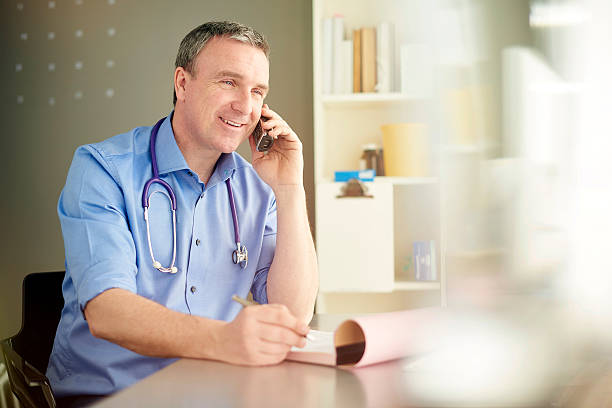 male gp gives good news over the phone male gp sits at his desk and speaks on the phone to his patient to check he's still alive . He's smiling so it must be good news . rolled up sleeves stock pictures, royalty-free photos & images