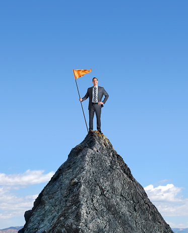 A confident businessman plants his flag on top of a mountain.  He smiles and looks directly at the camera as he holds the flagpole in one hand while placing his other hand on his hip.  A deep blue sky is behid him as it contrasts against the orange flag.