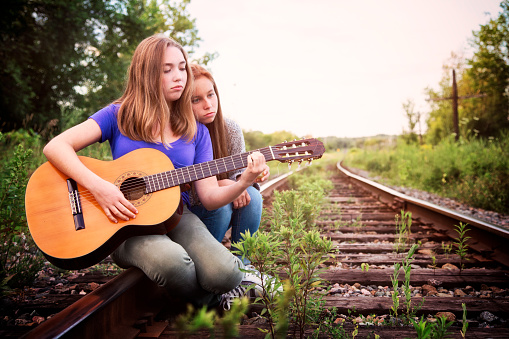 Heartbroken teenager plays sad song she wrote to friend on guitar. They are both siting outdoors on a railroad track.