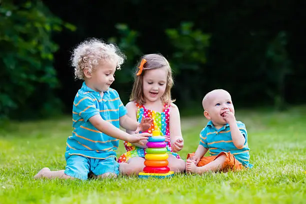 Three little children play with colorful rainbow pyramid toy. Educational toys for young child. Sibling kids building tower together. Toddler boy, preschooler girl and baby build blocks outdoors.