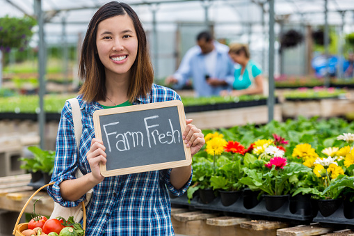 Beautiful young Vietnames woman holds a basket full of fresh veggies and a chalkboard 'Farm Fresh' sign. Fowers and plants are in the background. People are also shopping in the background.