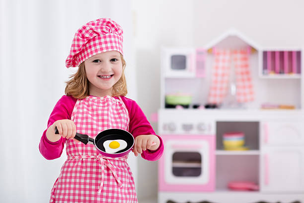 Little girl in chef hat and apron cooking fried eggs in toy kitchen.  Wooden toys for young children. Kids play and cook at home or daycare. Toddler kid playing with stove, tableware, pans and dishes.
